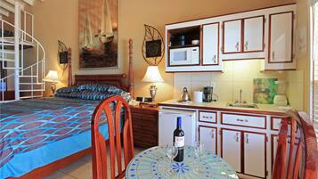 Quarto com cama, uma cadeira, mesa com garrafa e copos, escrivaninha com abajur e cozinha compacta com armários e micro-ondas.