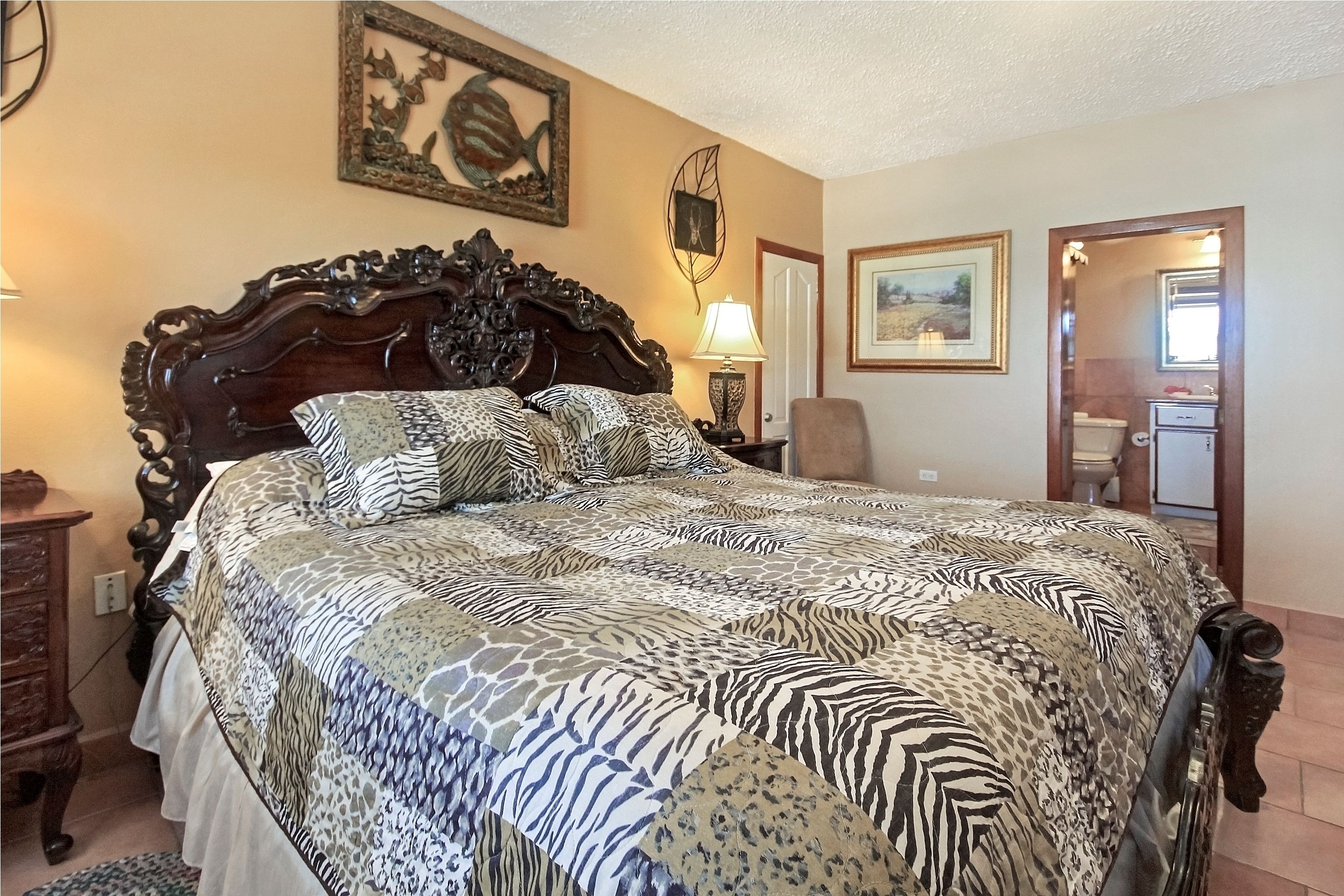 Une chambre avec un grand lit, une tête de lit en bois, une table de chevet avec une lampe, une chaise et une porte donnant sur une salle de bain.