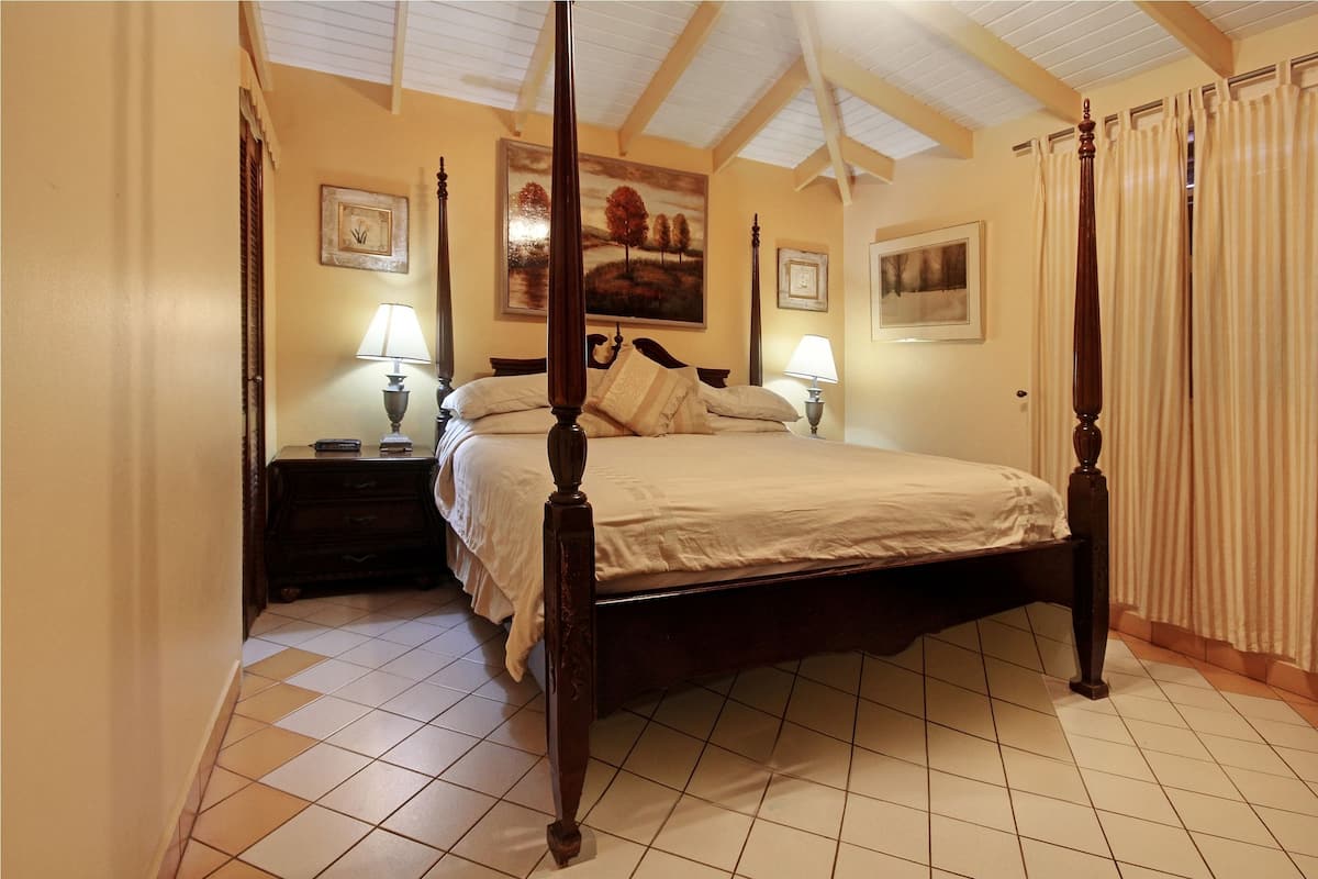 Uma cama com dossel de madeira, uma mesa de cabeceira com abajur e um quadro na parede.