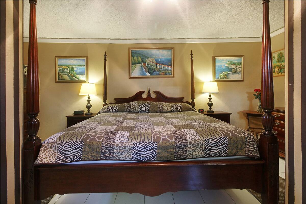 Uma cama de dossel com colcha estampada, duas luminárias de cabeceira e três quadros de paisagens nas paredes.