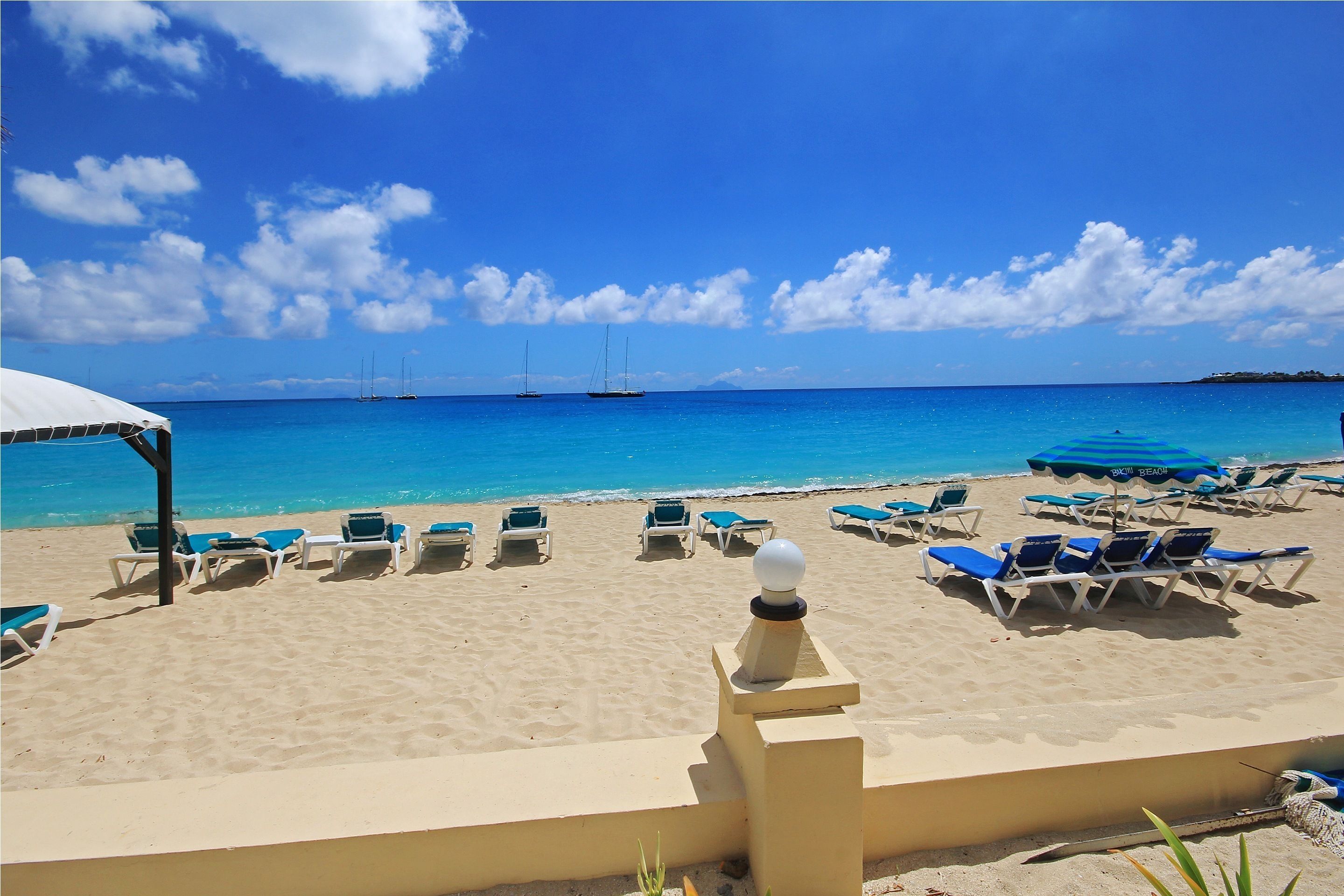 A beach with lounge chairs and umbrellas, a clear blue sky, and sailboats in the distance.