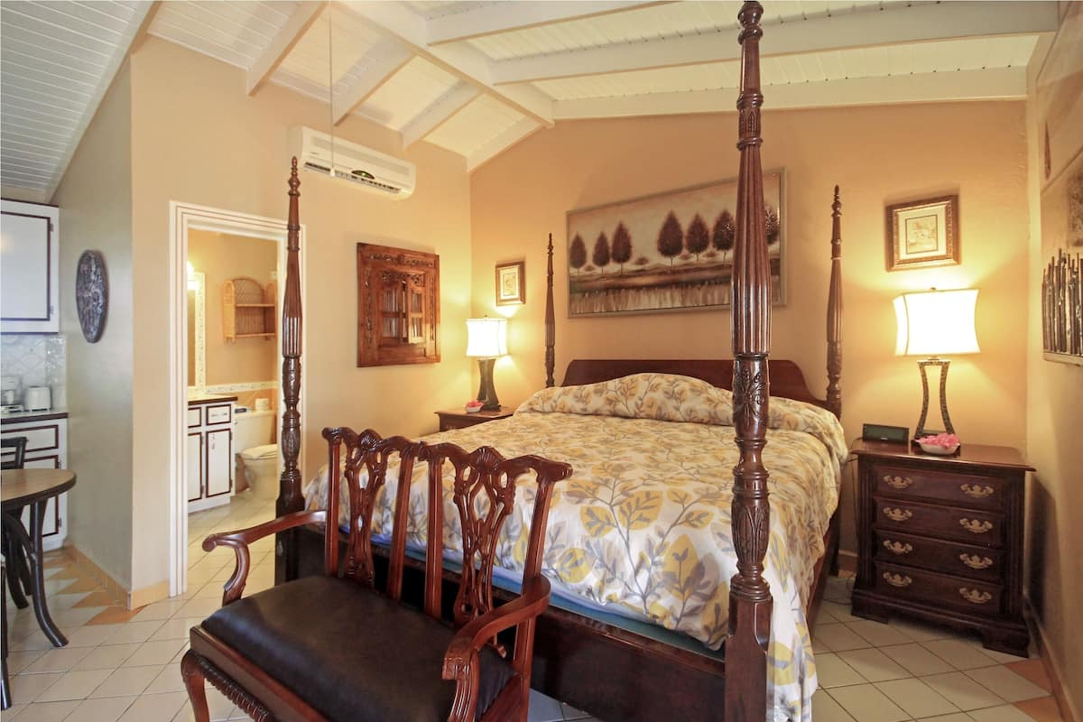 Uma cama com dossel com colcha florida, mesas de cabeceira de madeira com luminárias, uma cadeira de madeira e uma pequena mesa com abajur.