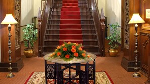 A grand staircase with a red carpet leading to an upper floor, flanked by ornate wooden doors and a table with a floral centerpiece. - WelcomHeritage Kasmanda Palace (Mussoorie)