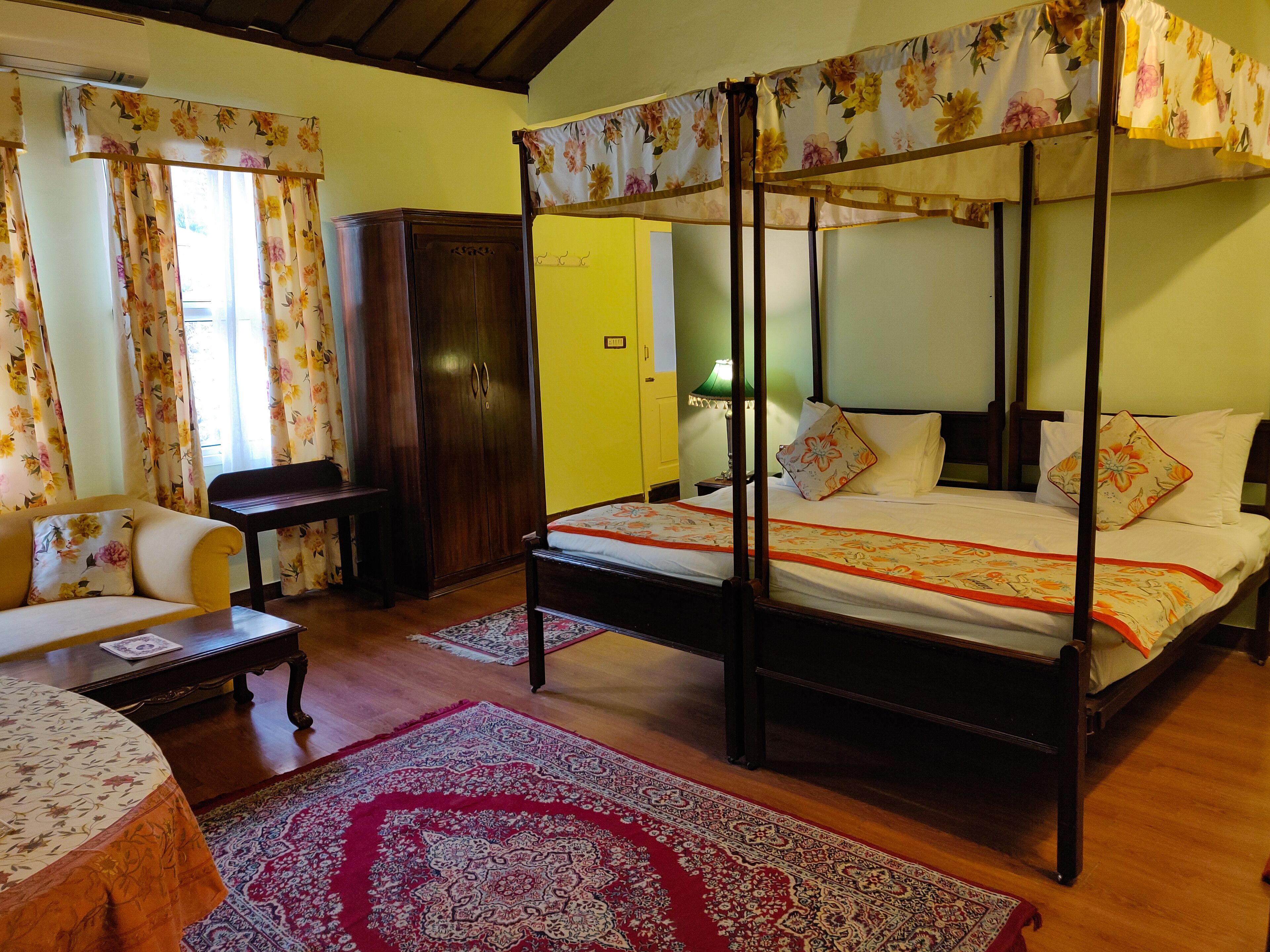 A four-poster bed with floral bedspread, a wooden wardrobe, a sofa with floral cushions, and a red patterned rug.