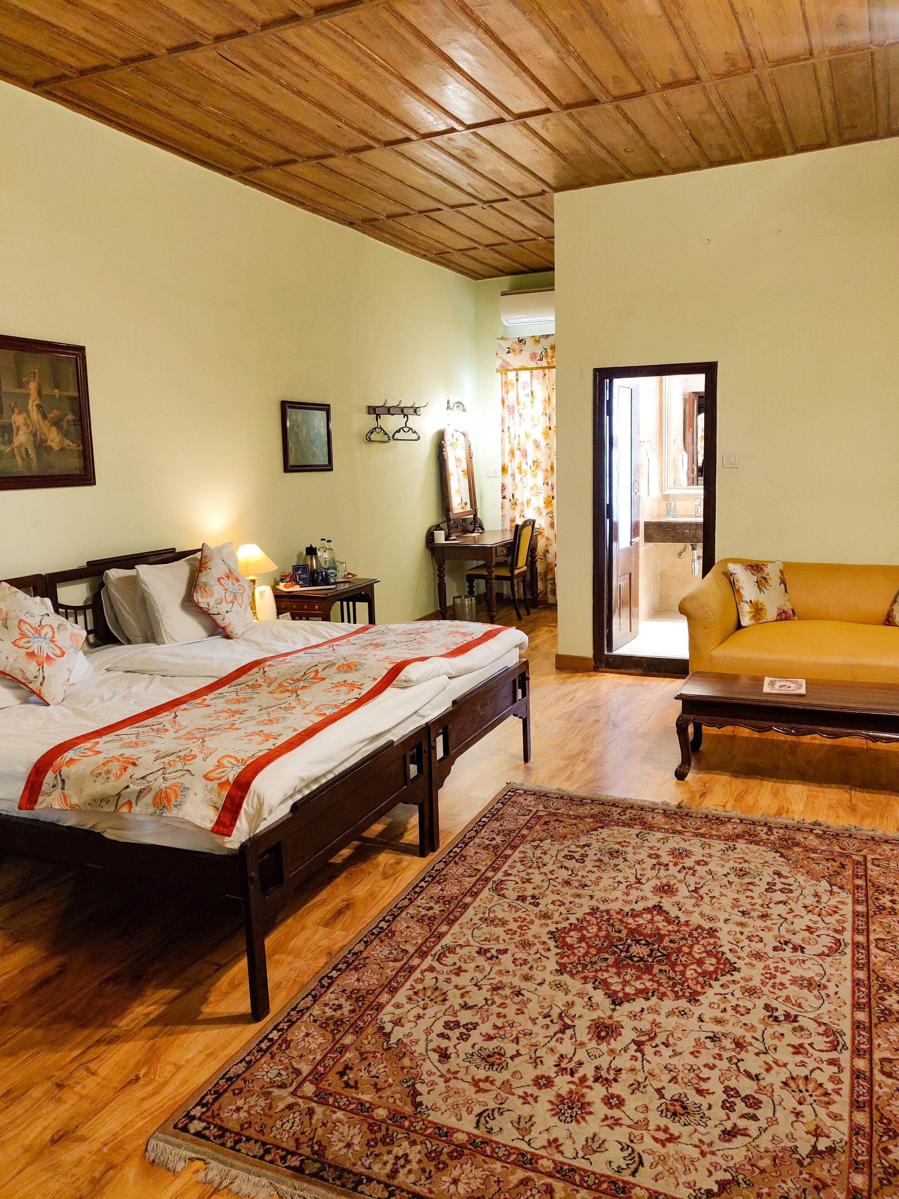 A bedroom with a wooden bed, a wooden desk, a mirror, a yellow sofa, and a patterned rug.