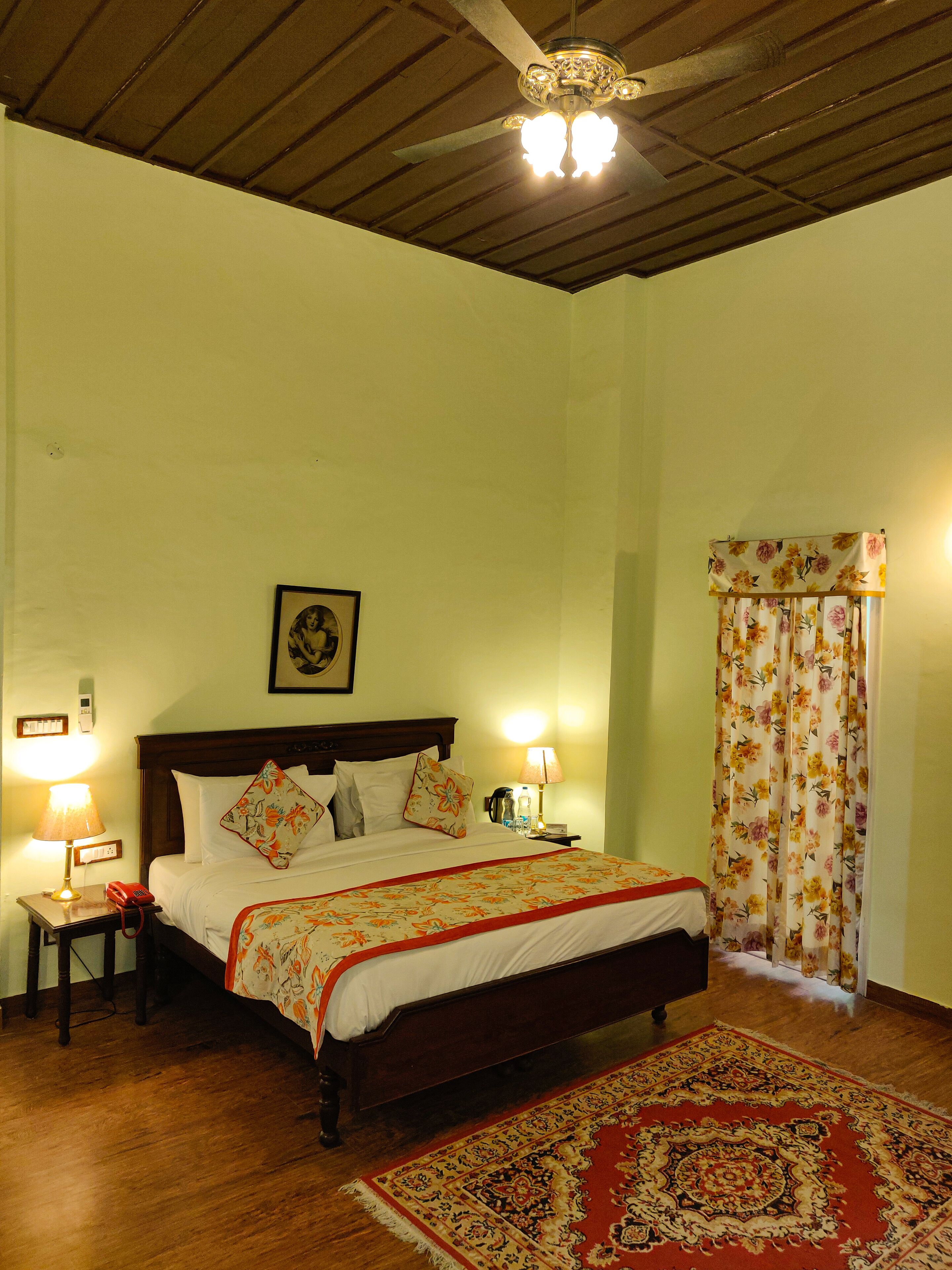 A bedroom with a wooden bed, two bedside lamps, a nightstand with a telephone, a framed picture on the wall, and a floral curtain.