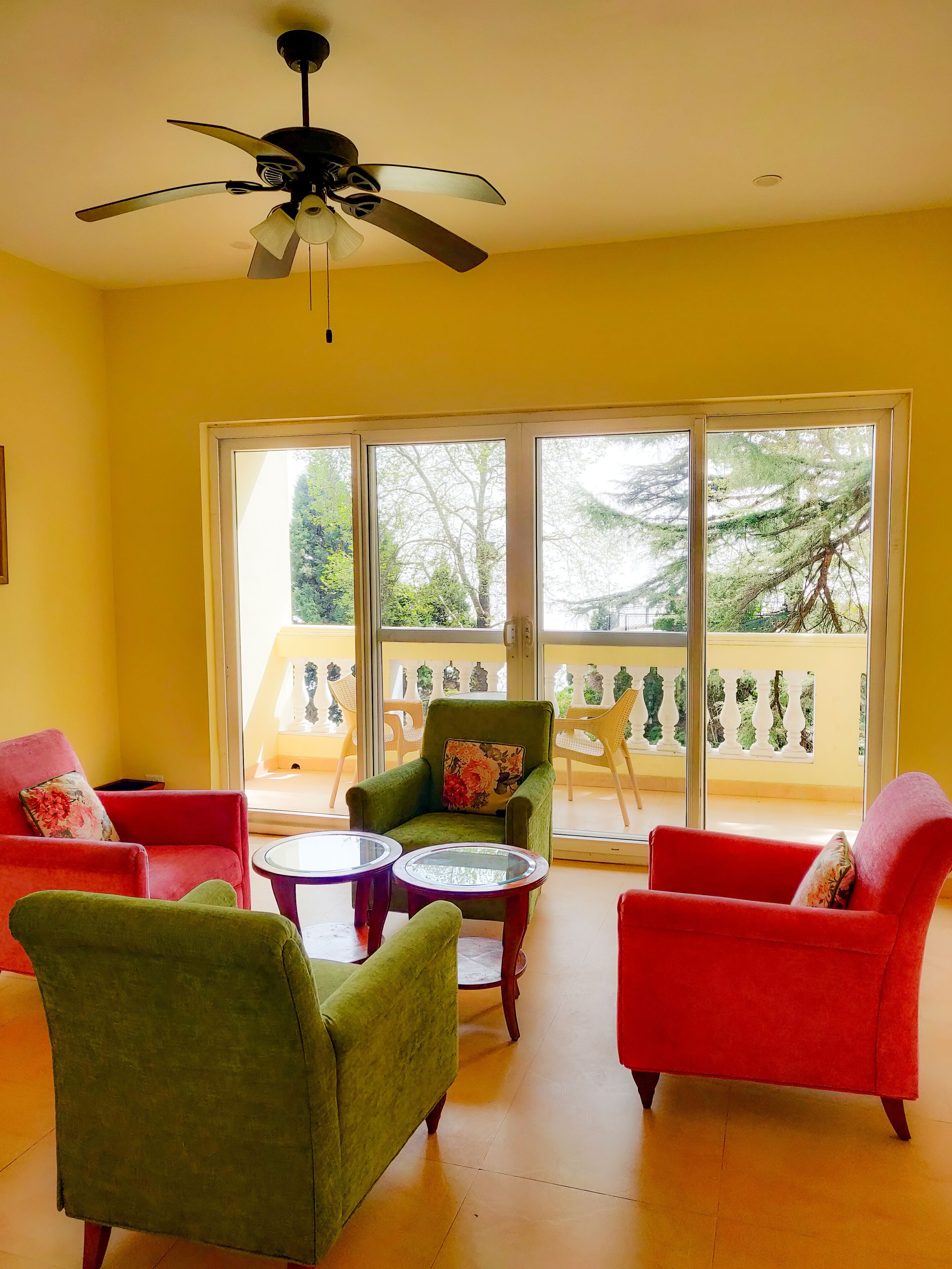 Una habitación con paredes amarillas, ventilador de techo, balcón con sillas y dos sillones rojos con cojines estampados.