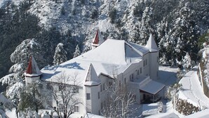 A snow-covered building with a symmetrical roof and two prominent spires, surrounded by snow-laden trees. - WelcomHeritage Kasmanda Palace (Mussoorie)