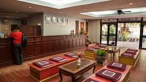 A hotel lobby with a reception desk, seating area with cushions, a coffee table, and a large window with a view of the outside.