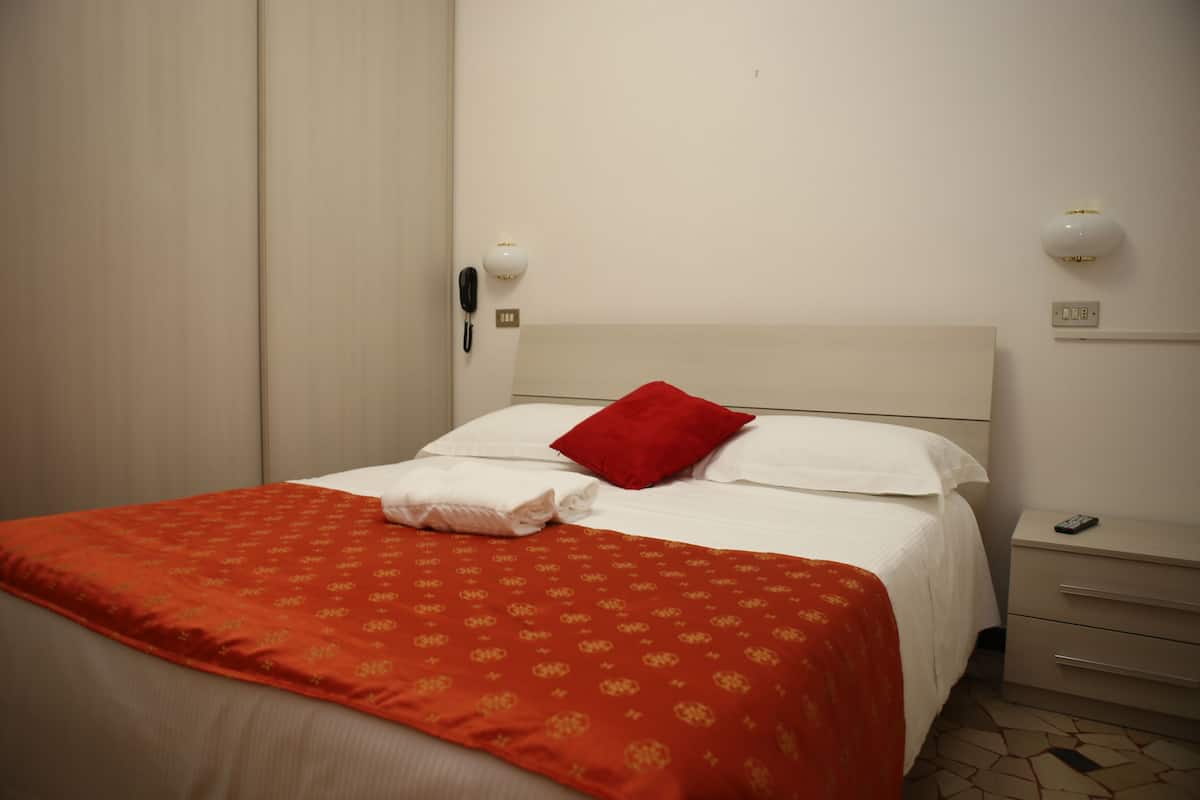 A hotel room with a neatly made bed, a red pillow, and a white blanket. There is a bedside table with a lamp and a phone.