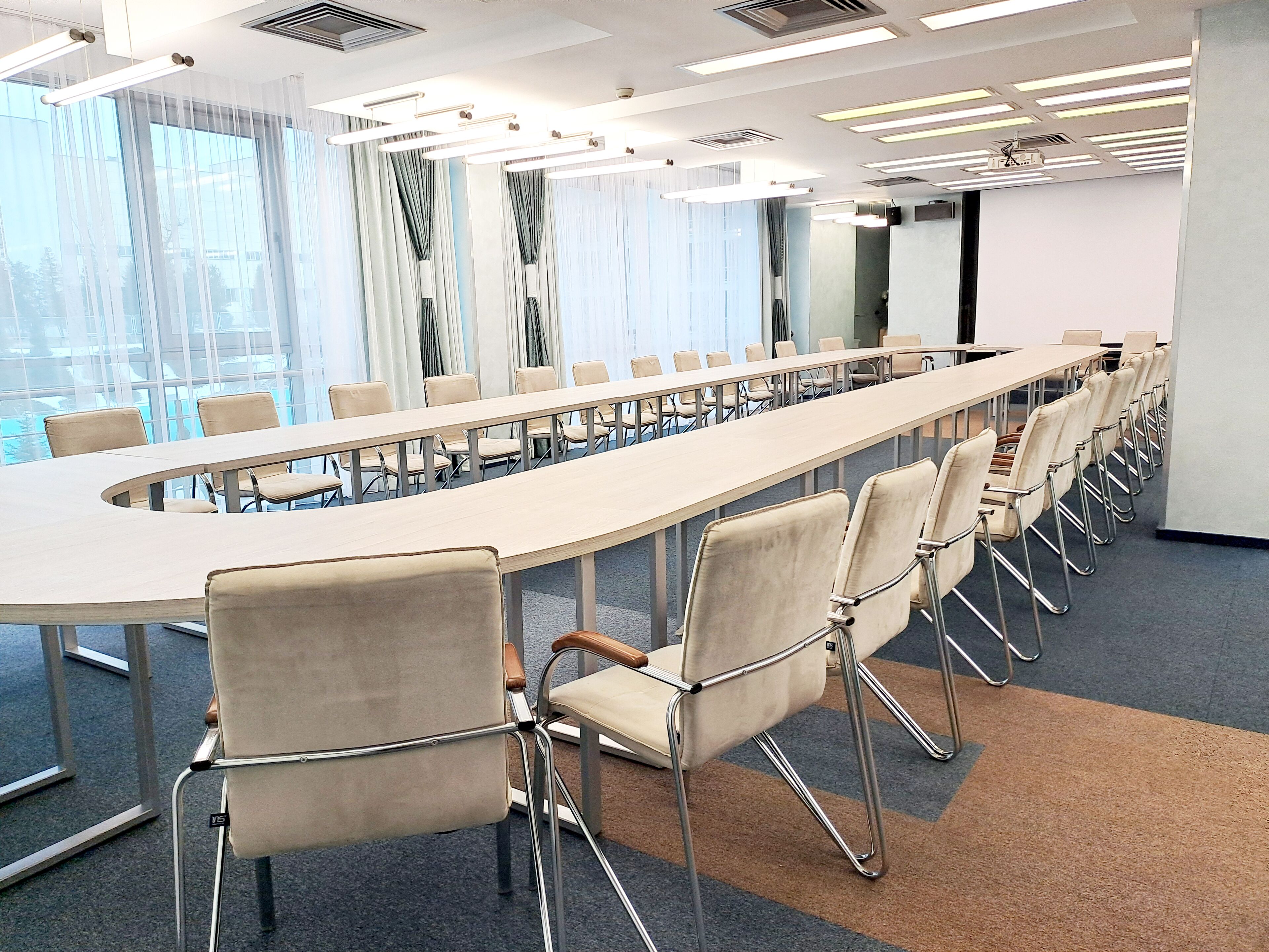 A conference room with a long table and chairs, large windows with curtains, and ceiling lights.