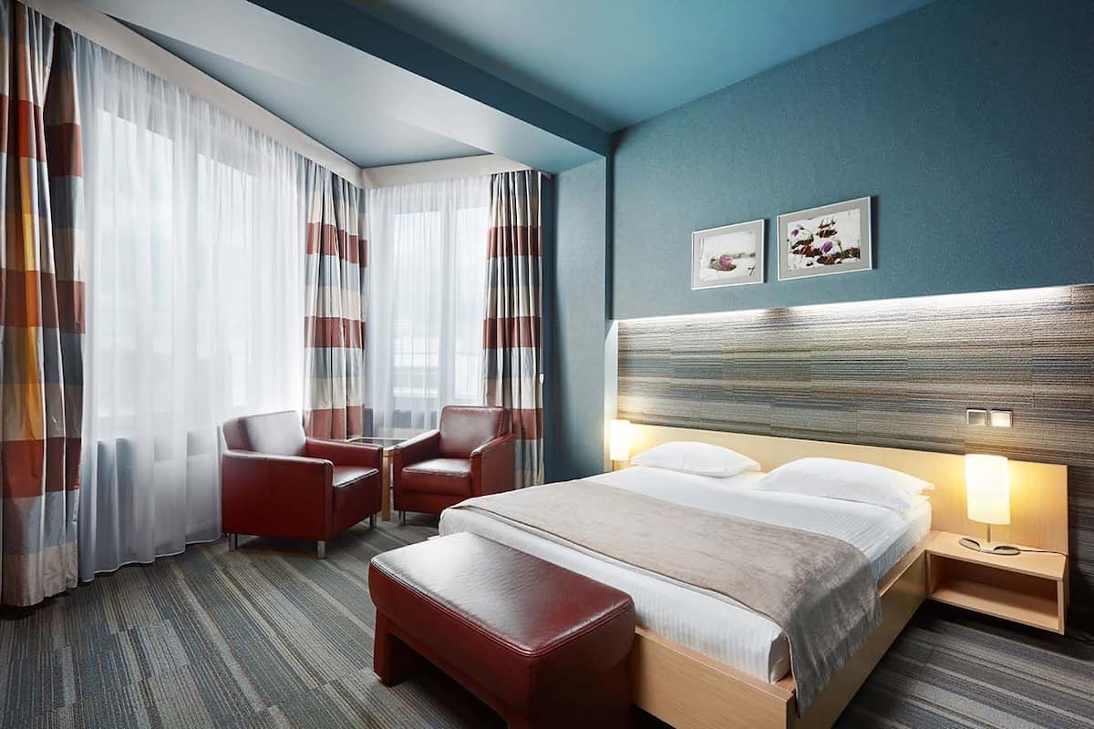 A hotel room with a bed, a desk, two chairs, a bench, and a nightstand with a lamp.