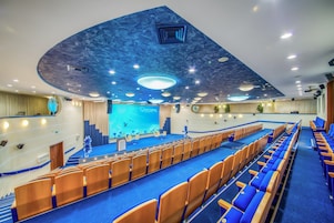 Un salón de conferencias con escenario, alfombra azul y filas de sillas.