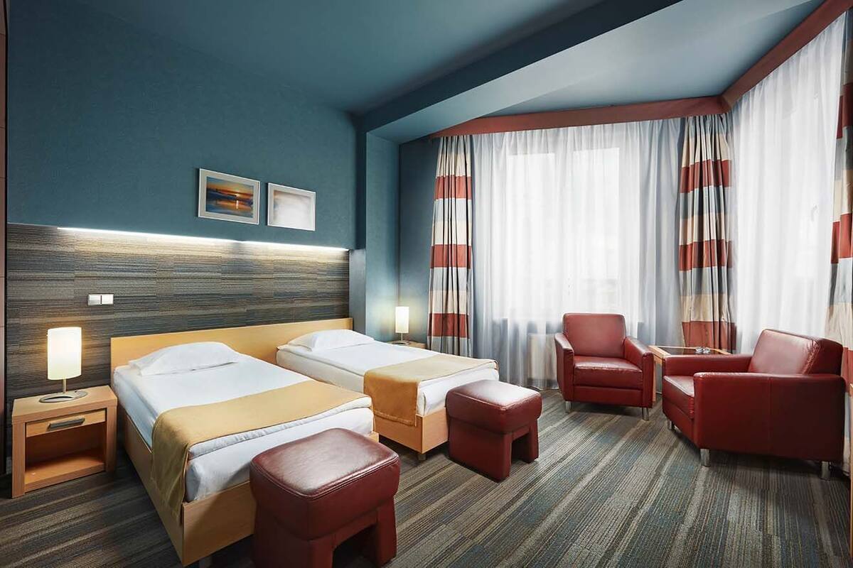 A hotel room with two beds, a red leather armchair, a wooden nightstand with a lamp, and a large window with curtains.