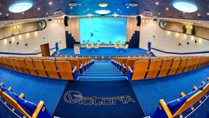 A large, circular auditorium with a stage at the front, rows of wooden and blue chairs, and a blue carpet with the Victoria logo. - Hotel Victoria Minsk (Minsk)