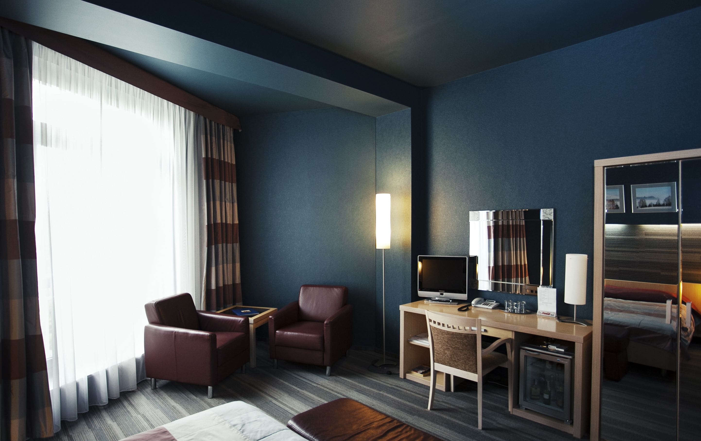 A hotel room with a desk, two chairs, a television, a lamp, a minibar, and a large mirror.