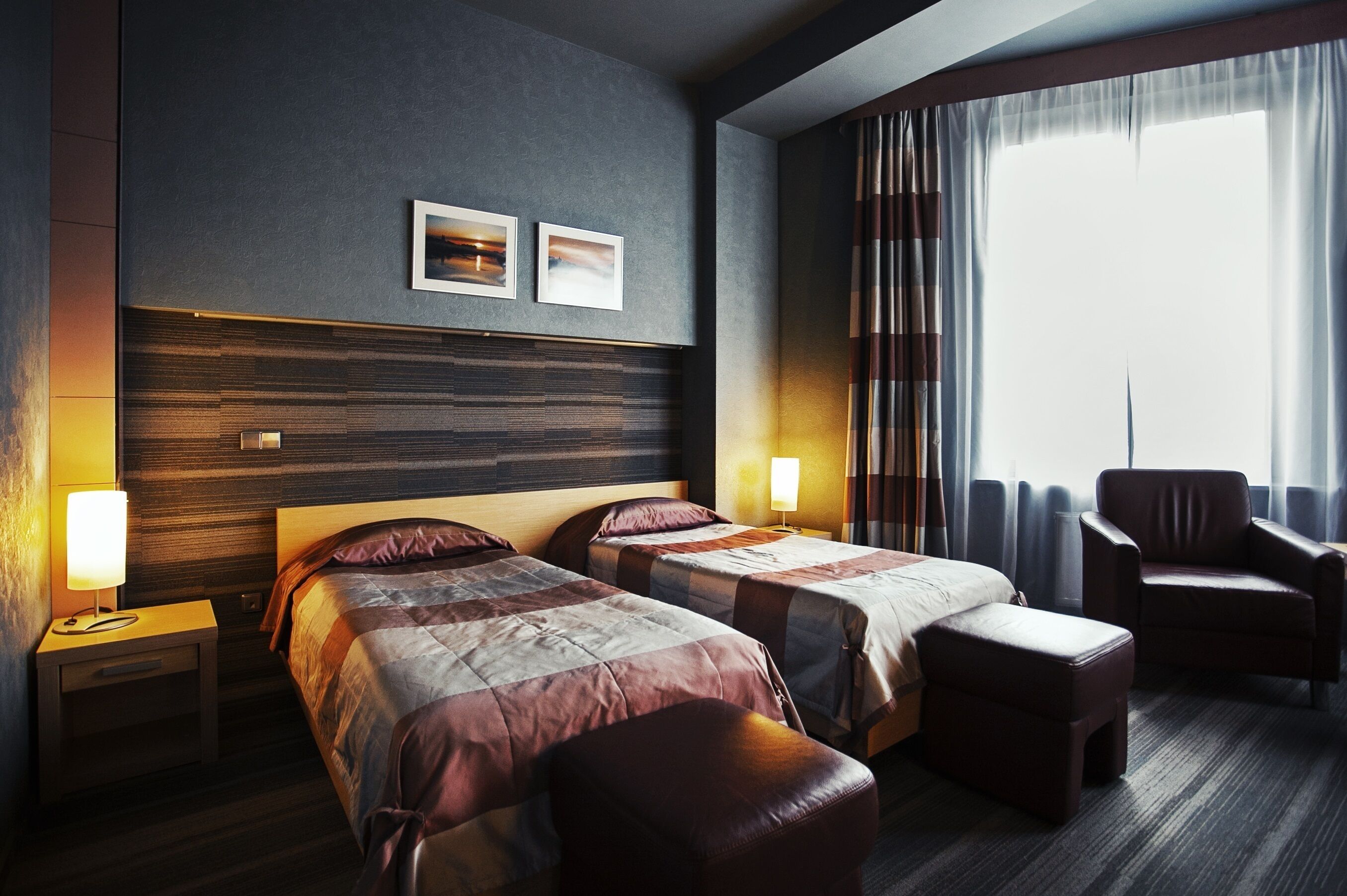 Habitación de hotel con dos camas, cabecero de madera, una silla y un ventanal con cortinas.