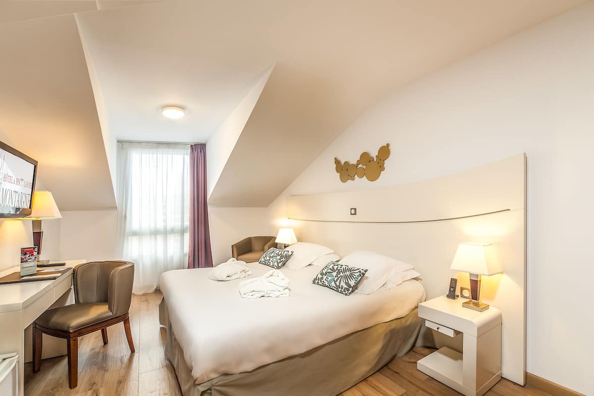 A hotel room with a bed, bedside tables, a desk, a chair, and a large window with curtains.