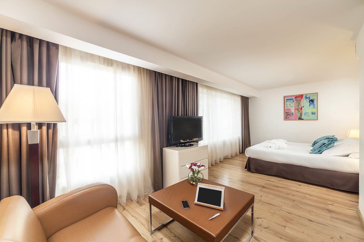 A hotel room with a bed, a TV, a desk with a lamp, a chair, and a window with curtains.