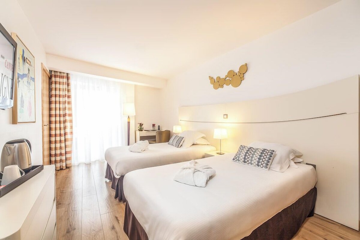 A hotel room with two beds, a desk, and a large window with curtains.