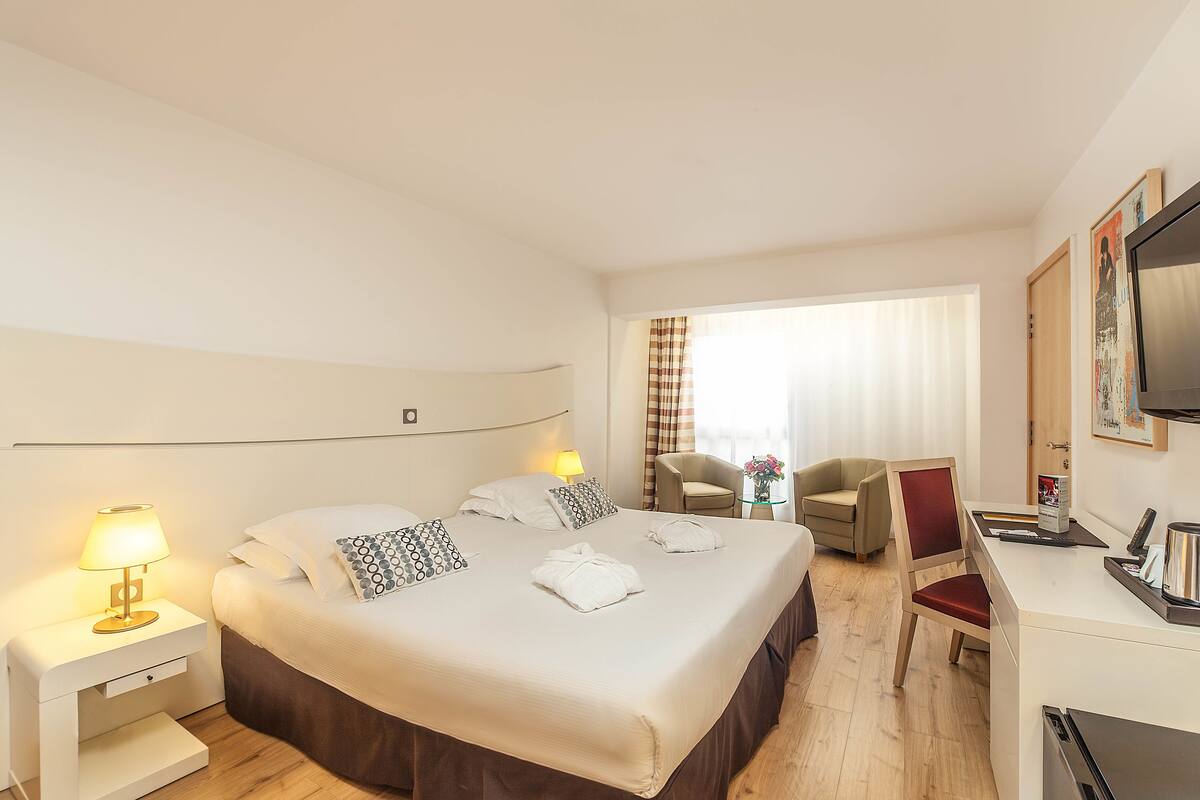 A hotel room with a large bed, bedside lamps, a desk with a chair, a television, and a window with curtains.