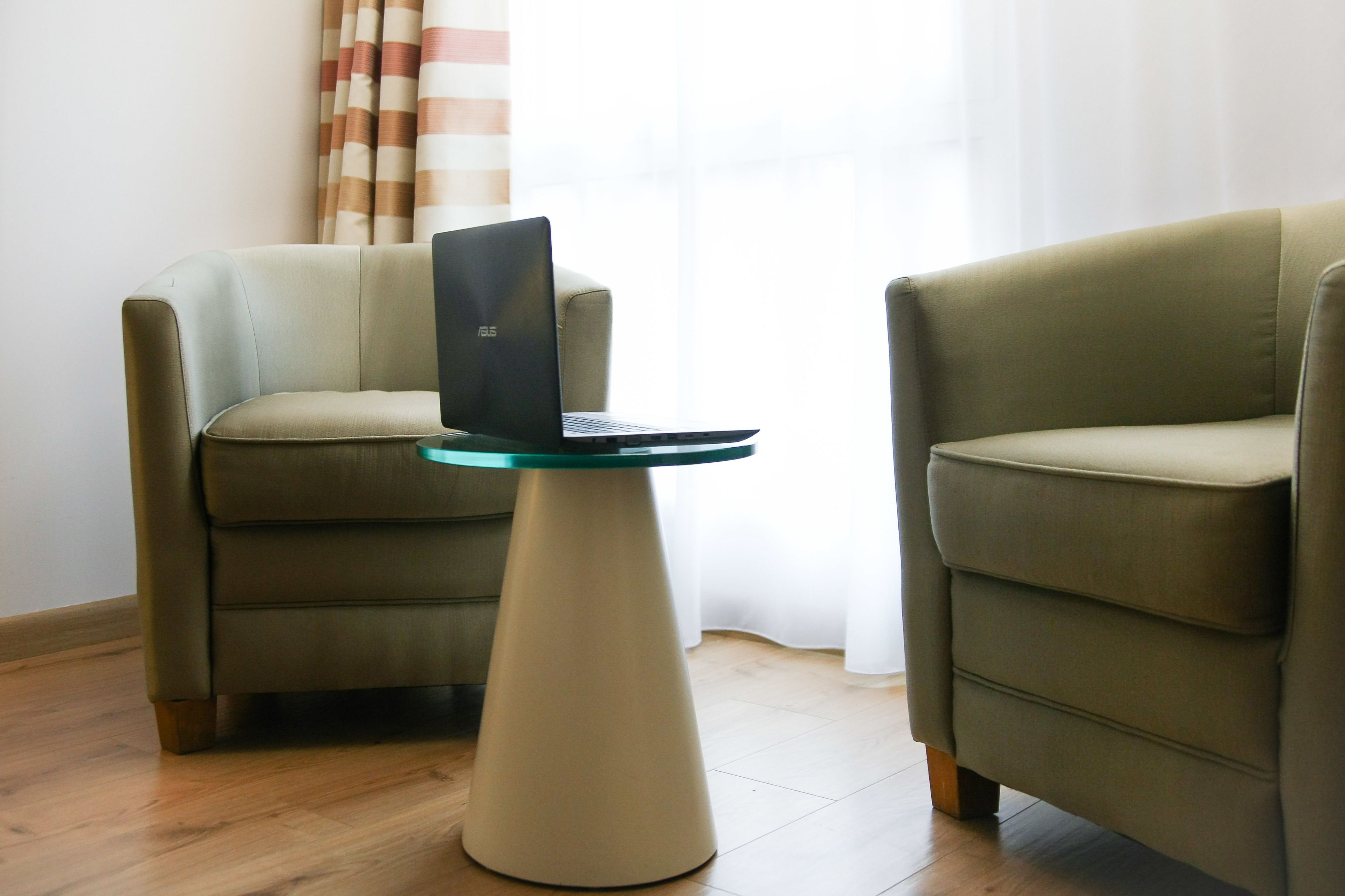 A room with two green armchairs, a small round table with a laptop, and a window with curtains.