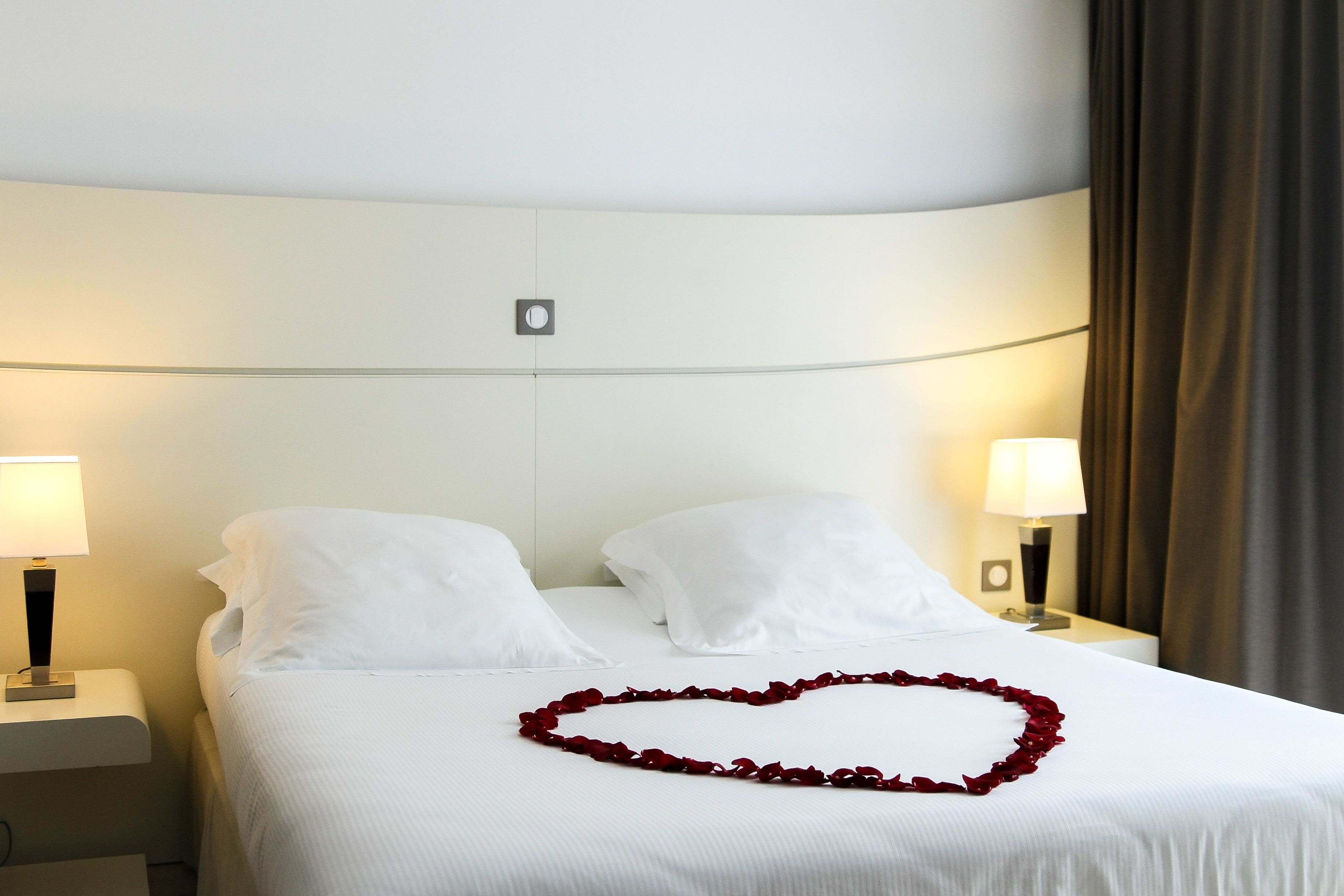 Una habitación de hotel con una cama bien hecha, dos lámparas de noche y una decoración con pétalos de rosa roja en forma de corazón.
