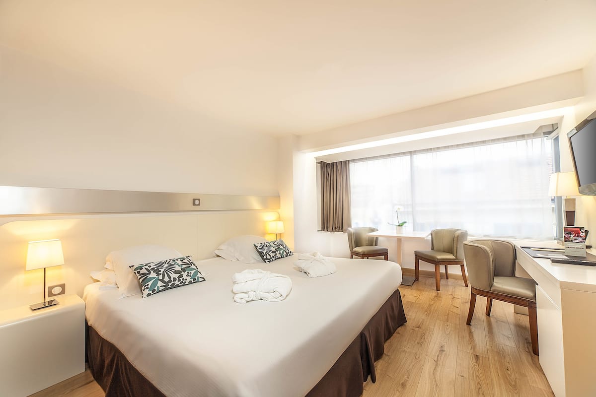 A hotel room with a large bed, two bedside tables with lamps, a desk with a chair, and a TV on the desk.