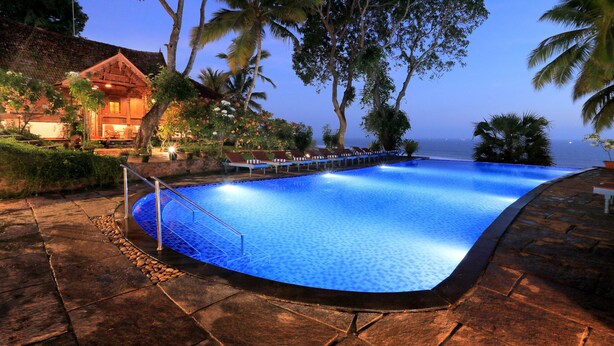 A swimming pool with a stone deck, surrounded by palm trees and a wooden building with a sloped roof. - Somatheeram Ayurveda Village (Neyyattinkara)