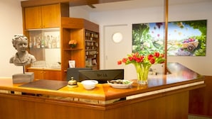 A reception desk with a wooden finish, a statue, a Dell computer, a vase with red flowers, a refrigerator with bottles, and a bookshelf. - Classic Hotel Kaarst (Kaarst)