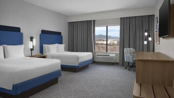 A hotel room with two beds, a desk, a chair, a TV, and a large window with a view of mountains.