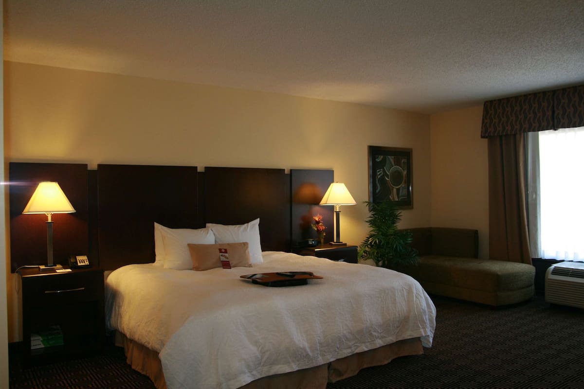 Ein Hotelzimmer mit einem großen Bett, zwei Nachttischlampen, einem kleinen Tisch, einem Sessel und einem Fenster mit Vorhängen.