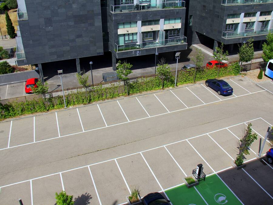A parking lot with multiple cars and a bus, surrounded by modern buildings with balconies and large windows.