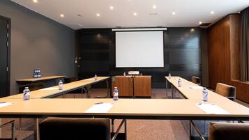 Une salle de conférence équipée de longues tables, de chaises, d’un écran de projection et d’un distributeur d’eau.