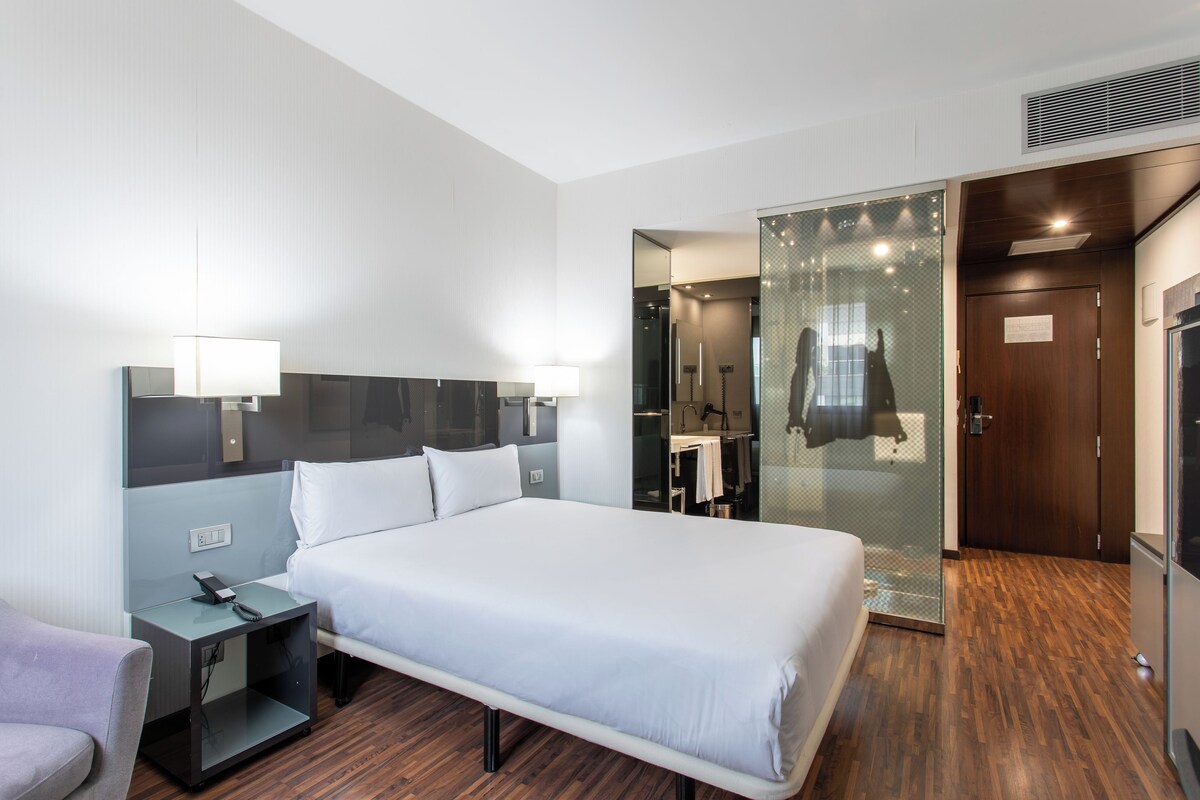 A hotel room with a large bed, bedside tables, a mirror, a wardrobe, and a shower area.