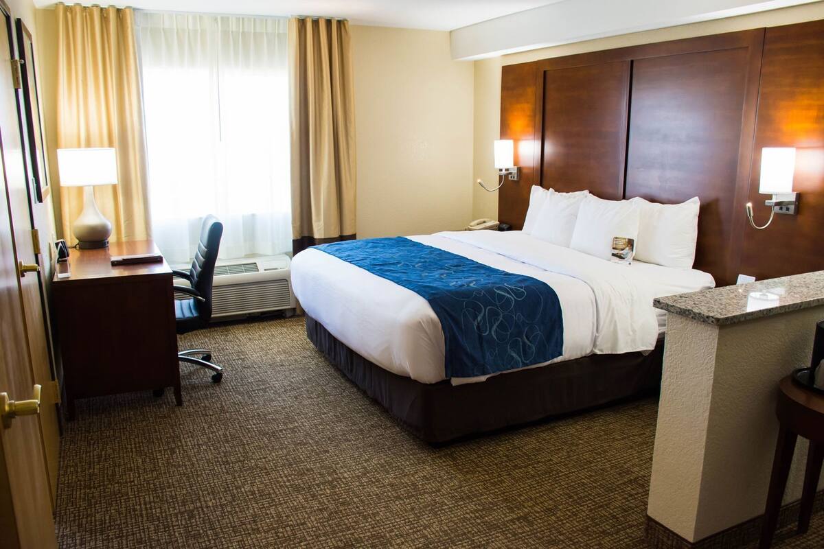 A hotel room with a bed, desk, chair, and a small kitchenette area.