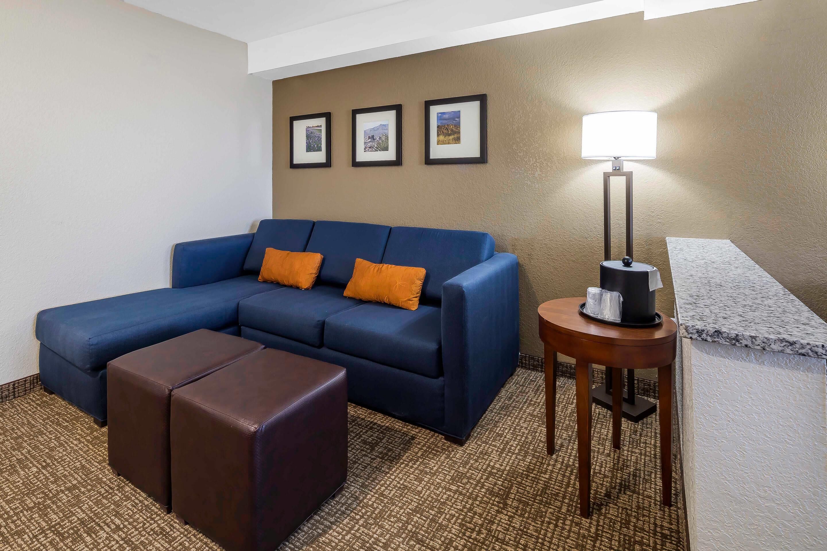 A hotel room with a blue sofa, two brown ottomans, a small wooden table with a lamp, and three framed pictures on the wall.