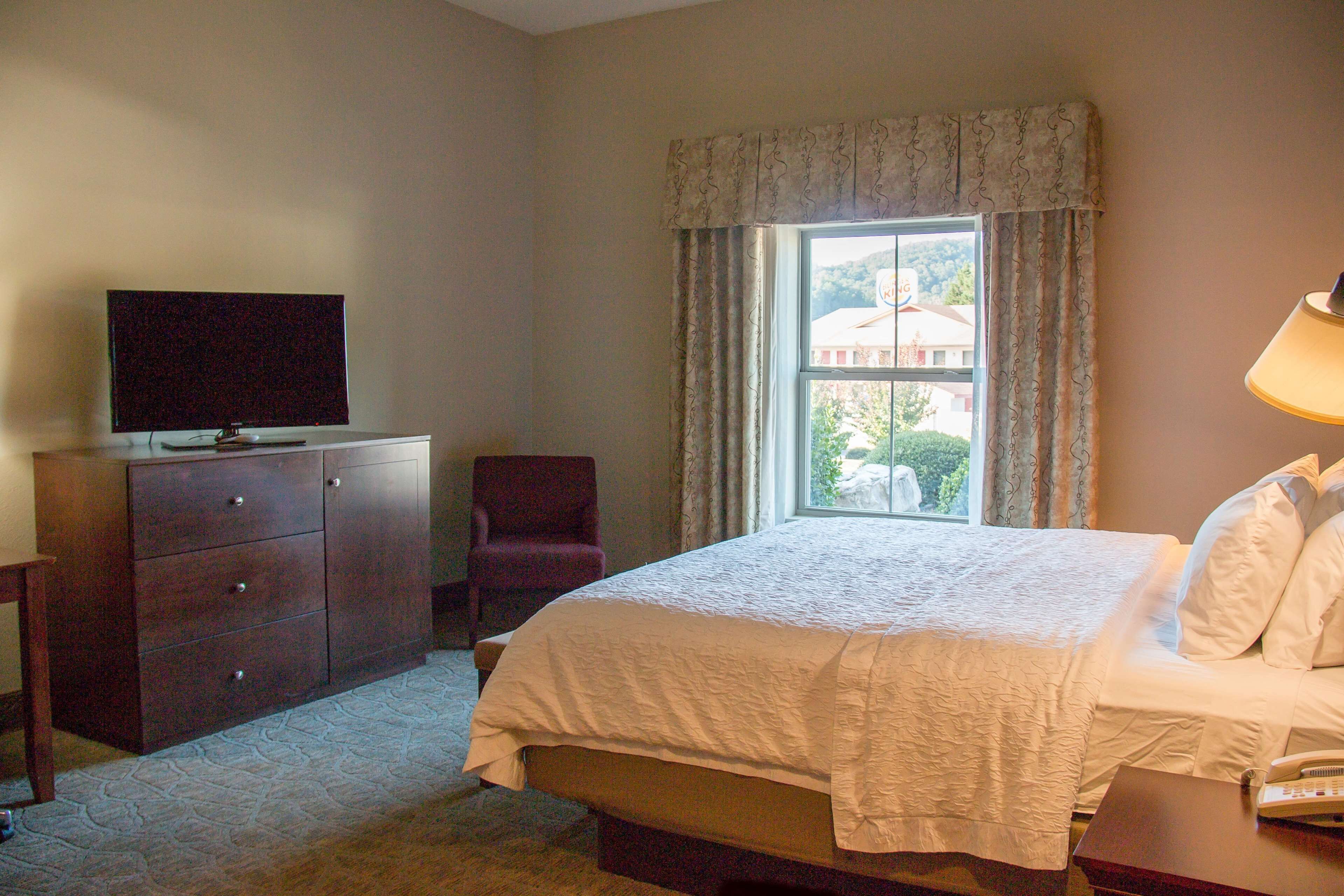 Quarto de hotel com uma cama, uma televisão num aparador, uma cadeira e uma janela com cortinas.
