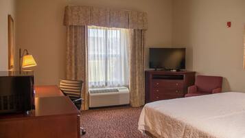 A hotel room with a bed, a desk with a lamp, a television, a chair, and an air conditioning unit.