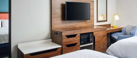 Une chambre d’hôtel moderne équipée d’un meuble TV en bois, d’un téléviseur à écran plat, d’un four à micro-ondes, d’une table de chevet et d’une chaise.