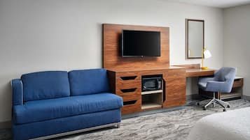 Une chambre d’hôtel avec un canapé bleu, un bureau en bois avec une télévision, une chaise et une petite lampe de bureau.