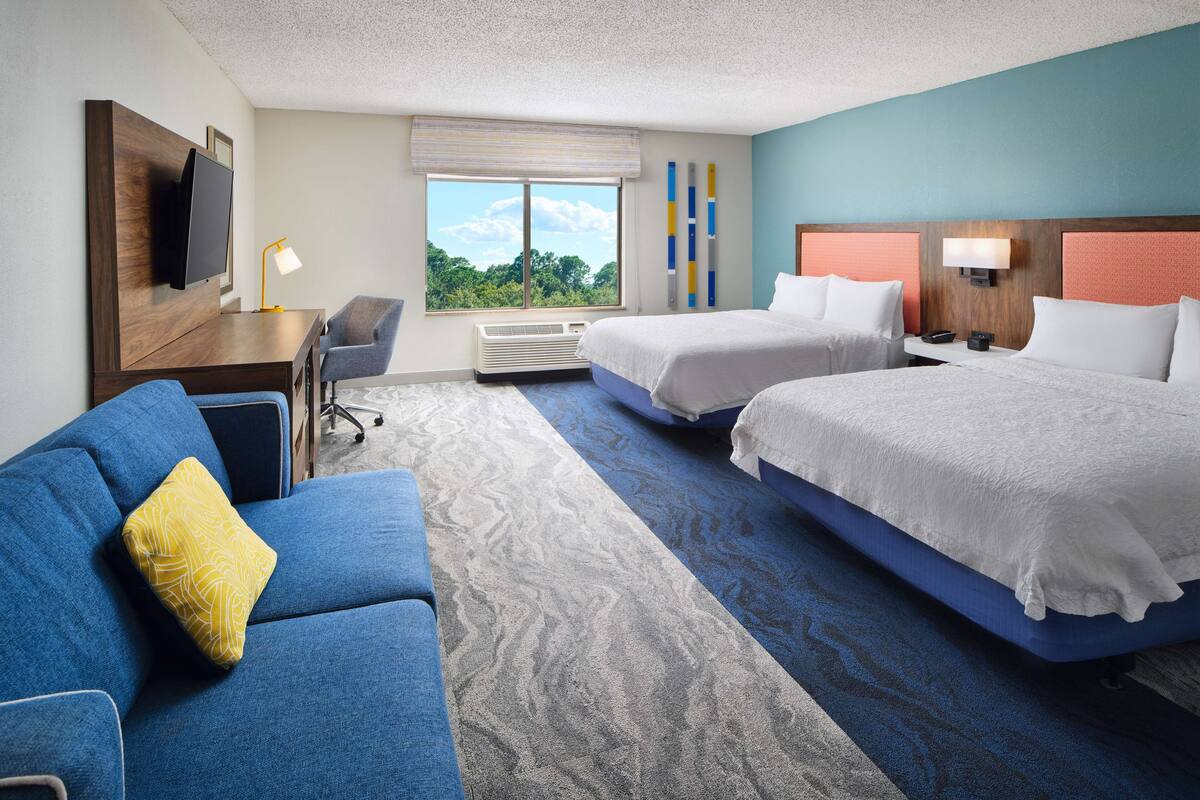 A hotel room with two beds, a sofa, a desk, a TV, and a window with a view of trees.