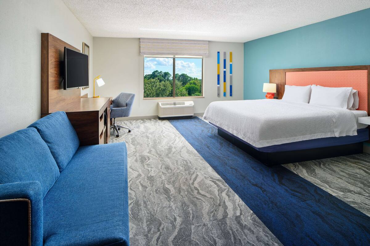 A hotel room with a bed, a sofa, a desk with a lamp, a TV, and a window with a view of trees.