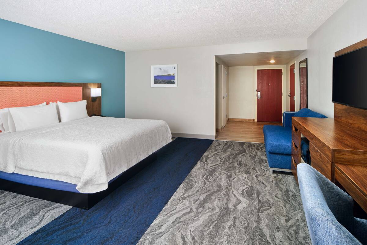 A hotel room with a bed, a desk, a chair, a TV, and a door leading to another room.