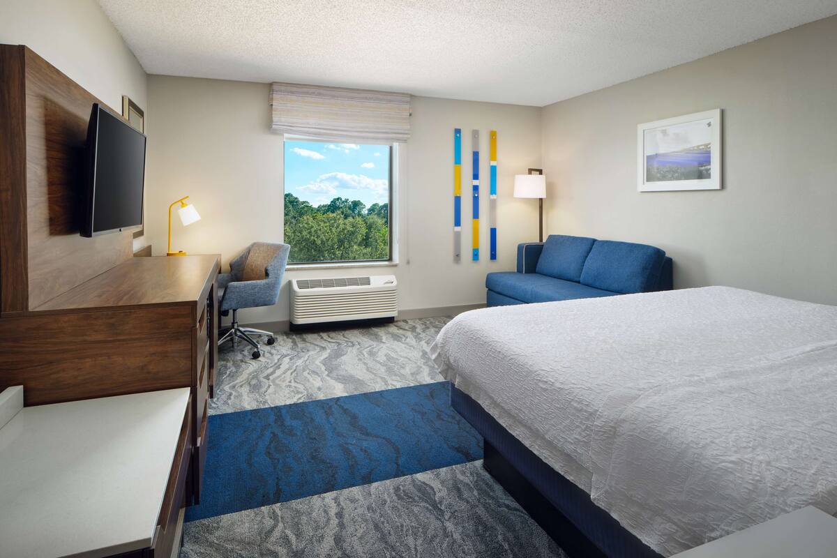 A hotel room with a bed, desk, chair, and a large window offering a view of greenery.