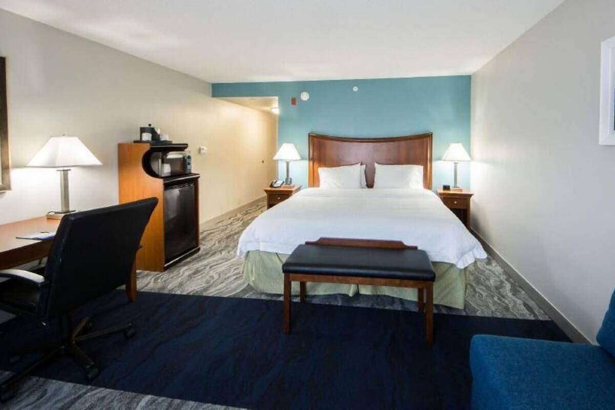 A hotel room with a bed, desk, chair, lamps, and a picture on the wall.