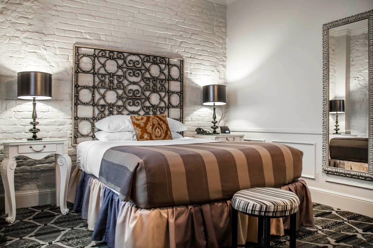 A bedroom with a patterned headboard, a bed with striped bedding, a bench, two bedside tables with lamps, and a large mirror.