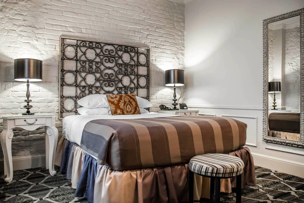 Un dormitorio con un cabecero estampado, una cama con ropa de cama a rayas, un banco, dos mesitas de noche con lámparas y un espejo grande.