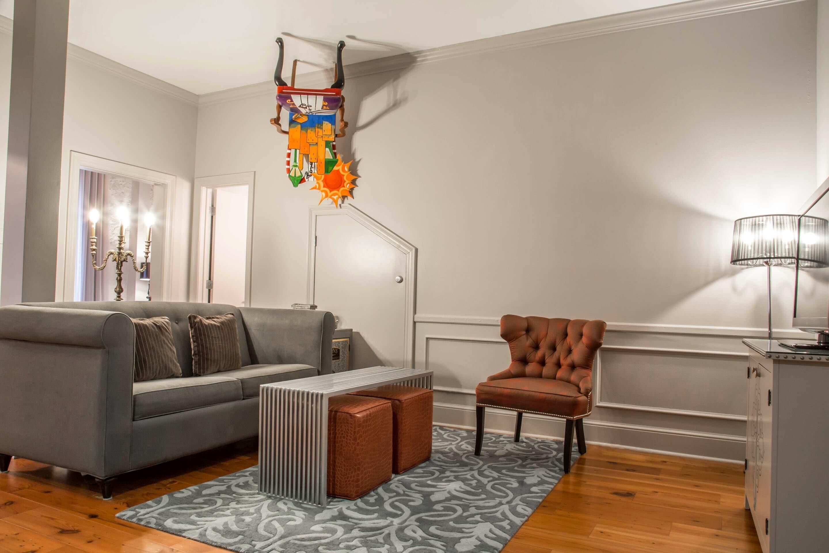 Un salon avec un canapé gris, un fauteuil marron, un tapis à motifs, une porte blanche et une étagère murale ornée d’objets décoratifs.