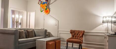 A living room with a grey sofa, a brown armchair, a patterned rug, a white door, and a wall-mounted shelf with decorative items.